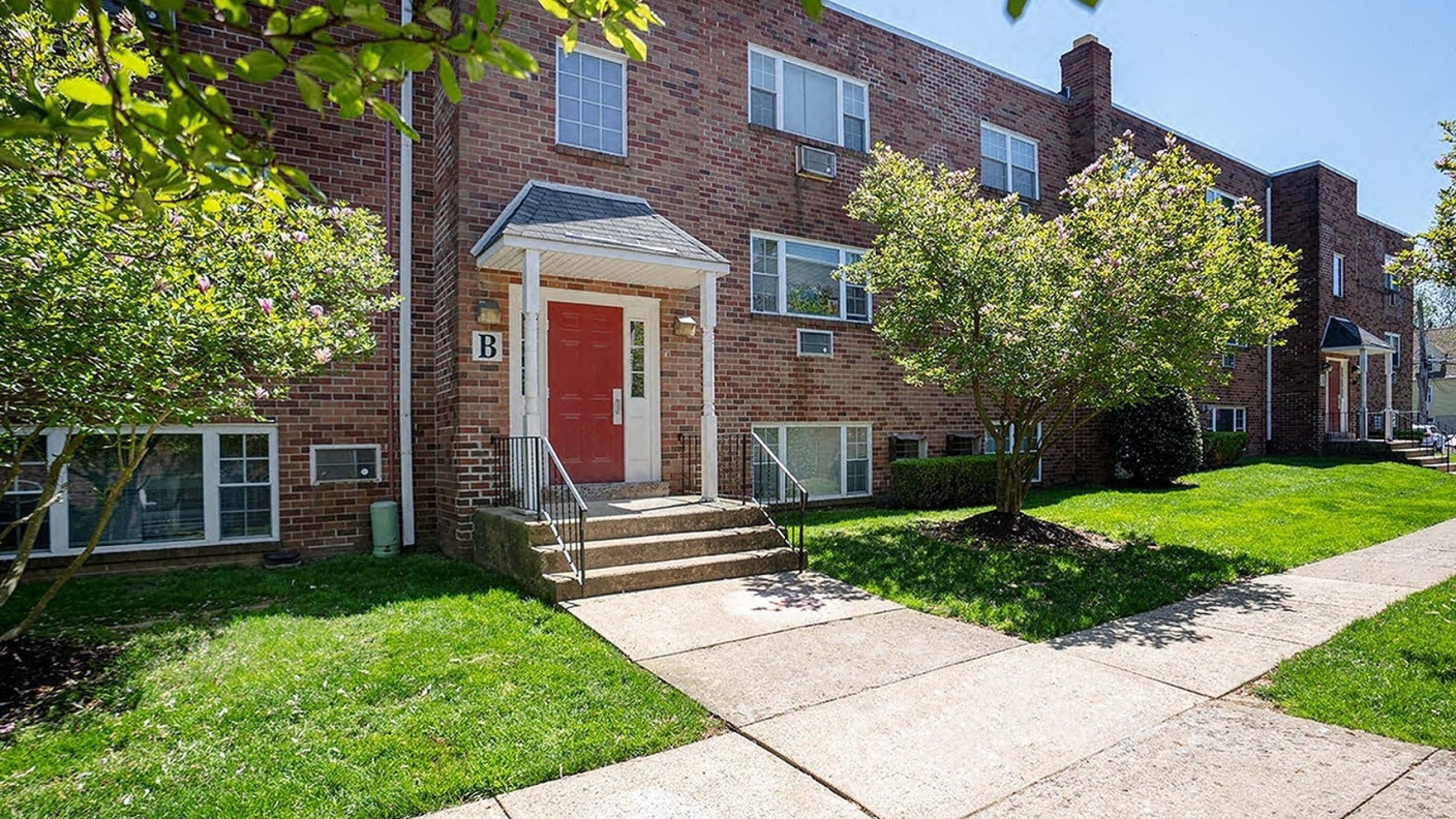 Charming brick apartment building with a welcoming entrance, lush greenery, and sunny surroundings.