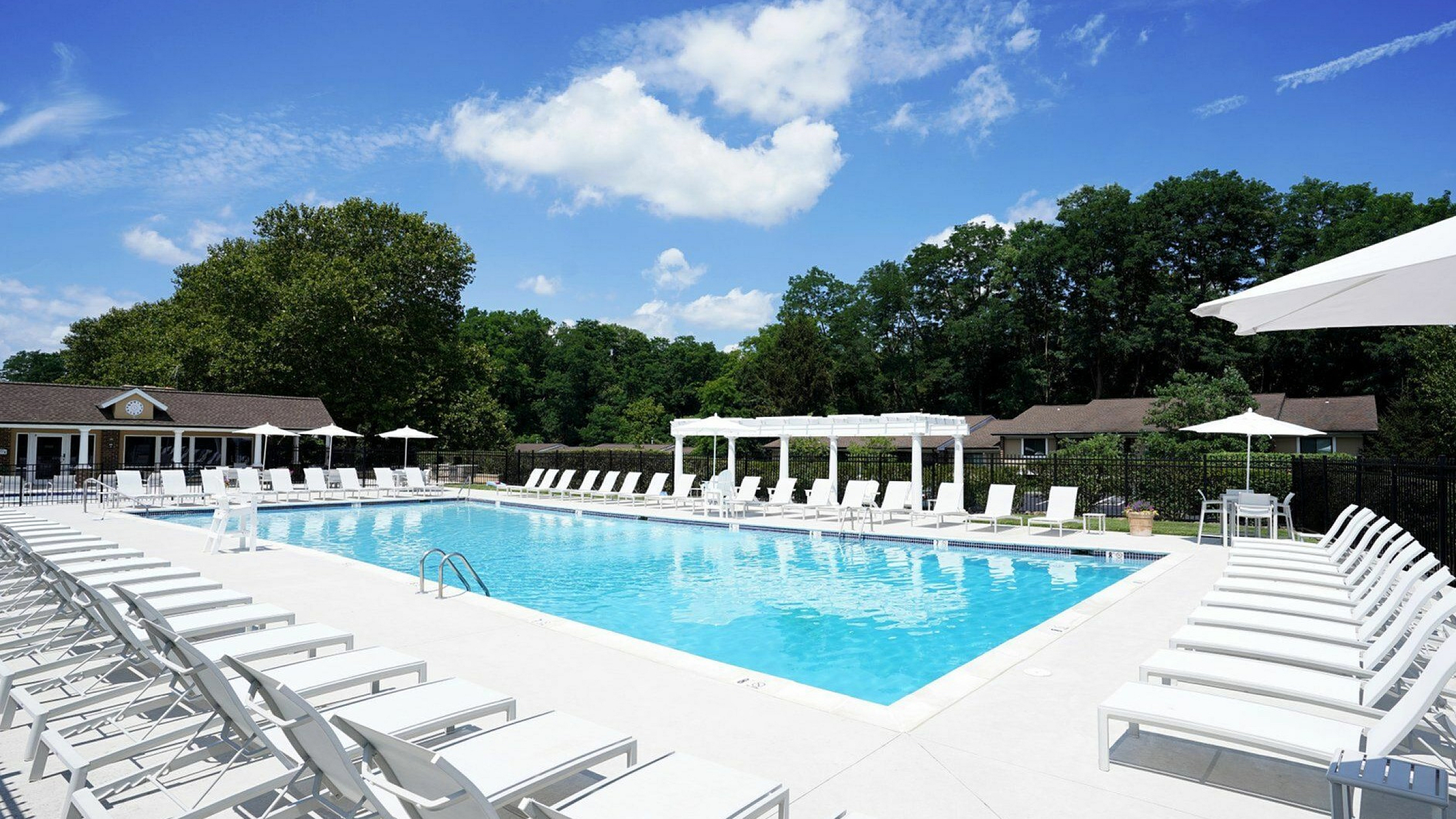 Elegant outdoor swimming pool area with abundant lounge chairs, surrounded by lush greenery and a bright blue sky, offering a perfect relaxation spot.