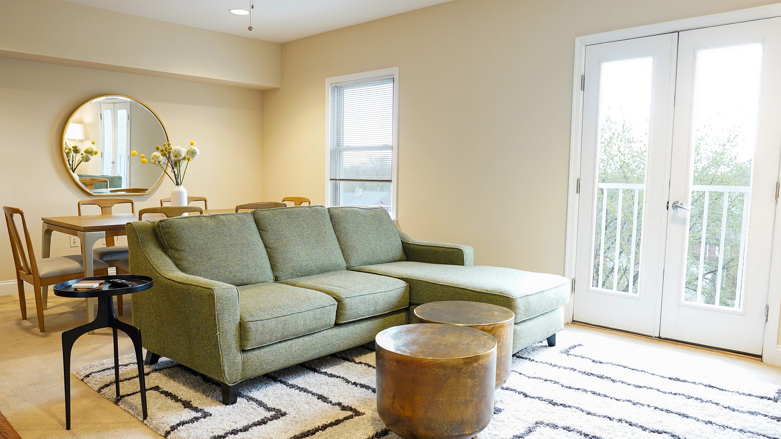Bright and cozy living room featuring a stylish green sectional sofa, elegant round coffee tables, and a chic dining area by the window. Natural light floods through large glass doors, highlighting the modern decor.