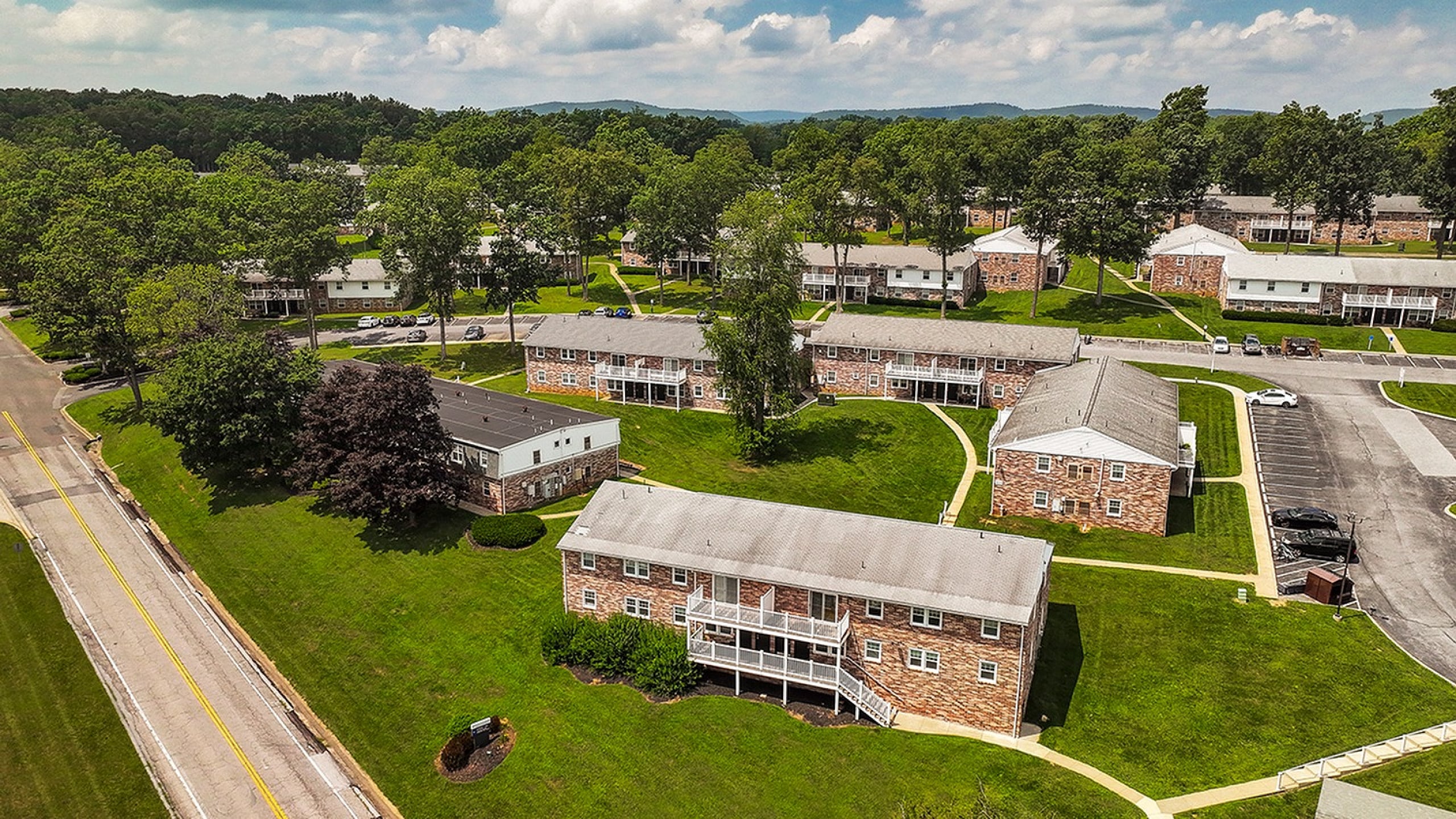 Charming aerial view of a beautifully landscaped apartment community with brick buildings, lush green lawns, and scenic surroundings.