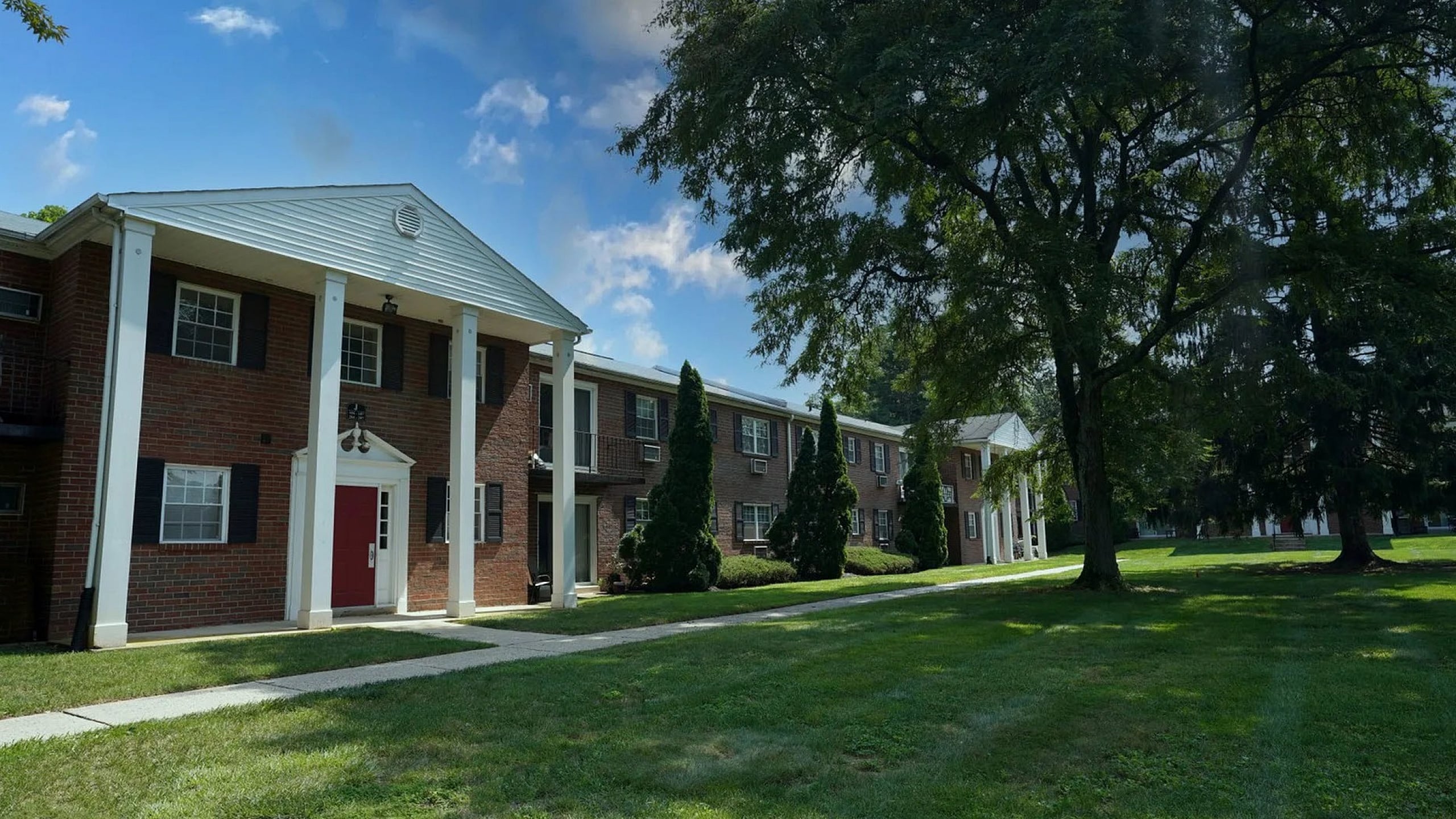 Charming red brick apartment complex with classic white column entrances and lush green lawns under a bright blue sky.
