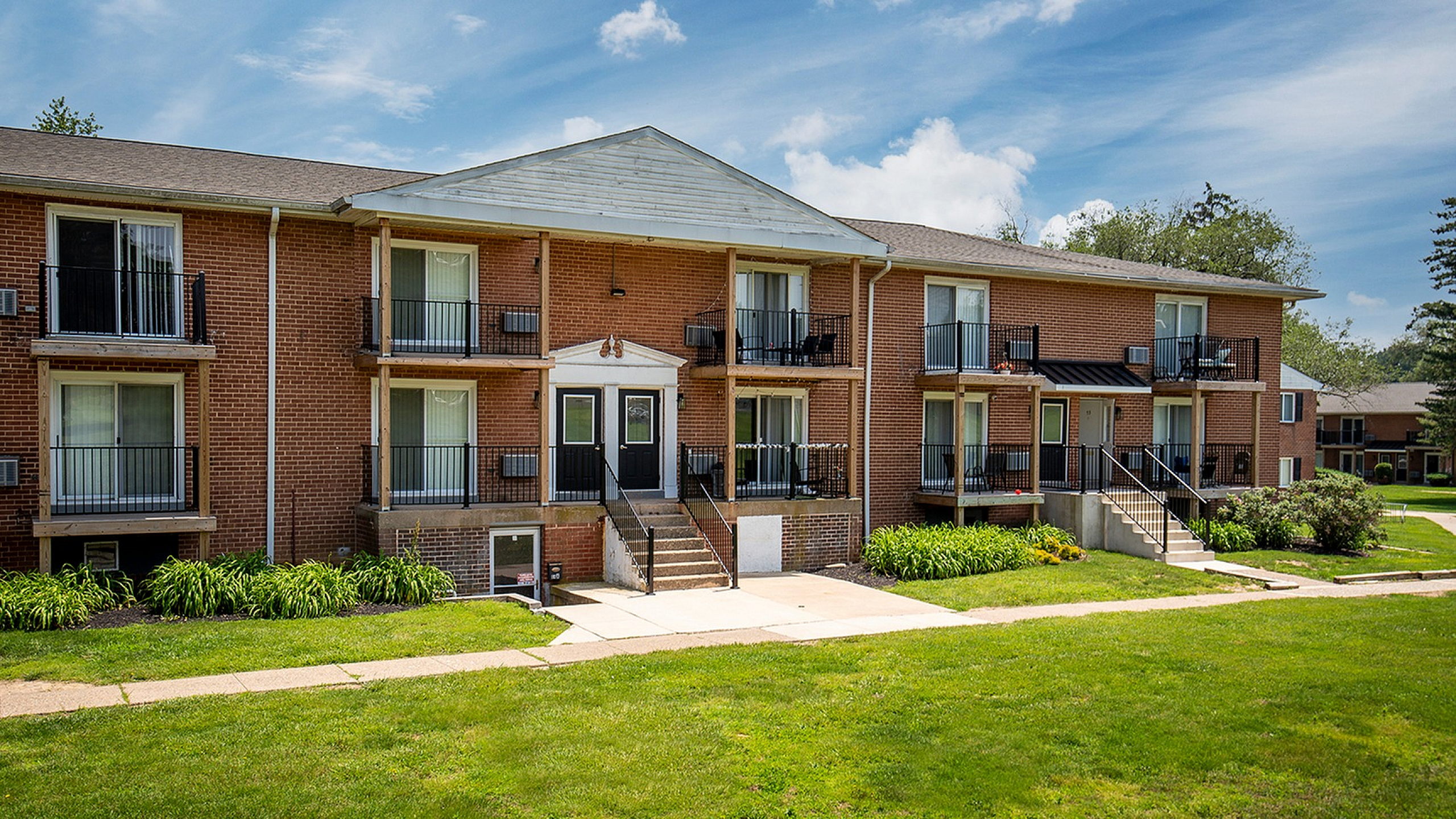 Charming apartment building with red brick facade, small balconies, and lush green lawns under a clear blue sky.