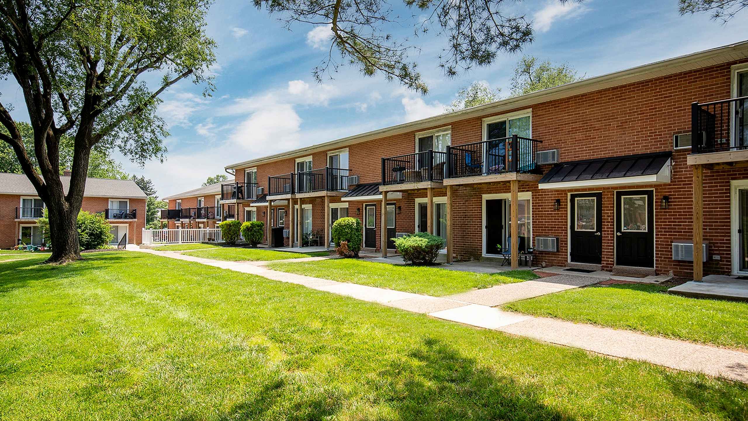 Charming brick apartments with private balconies and a lush green courtyard under a sunny sky.