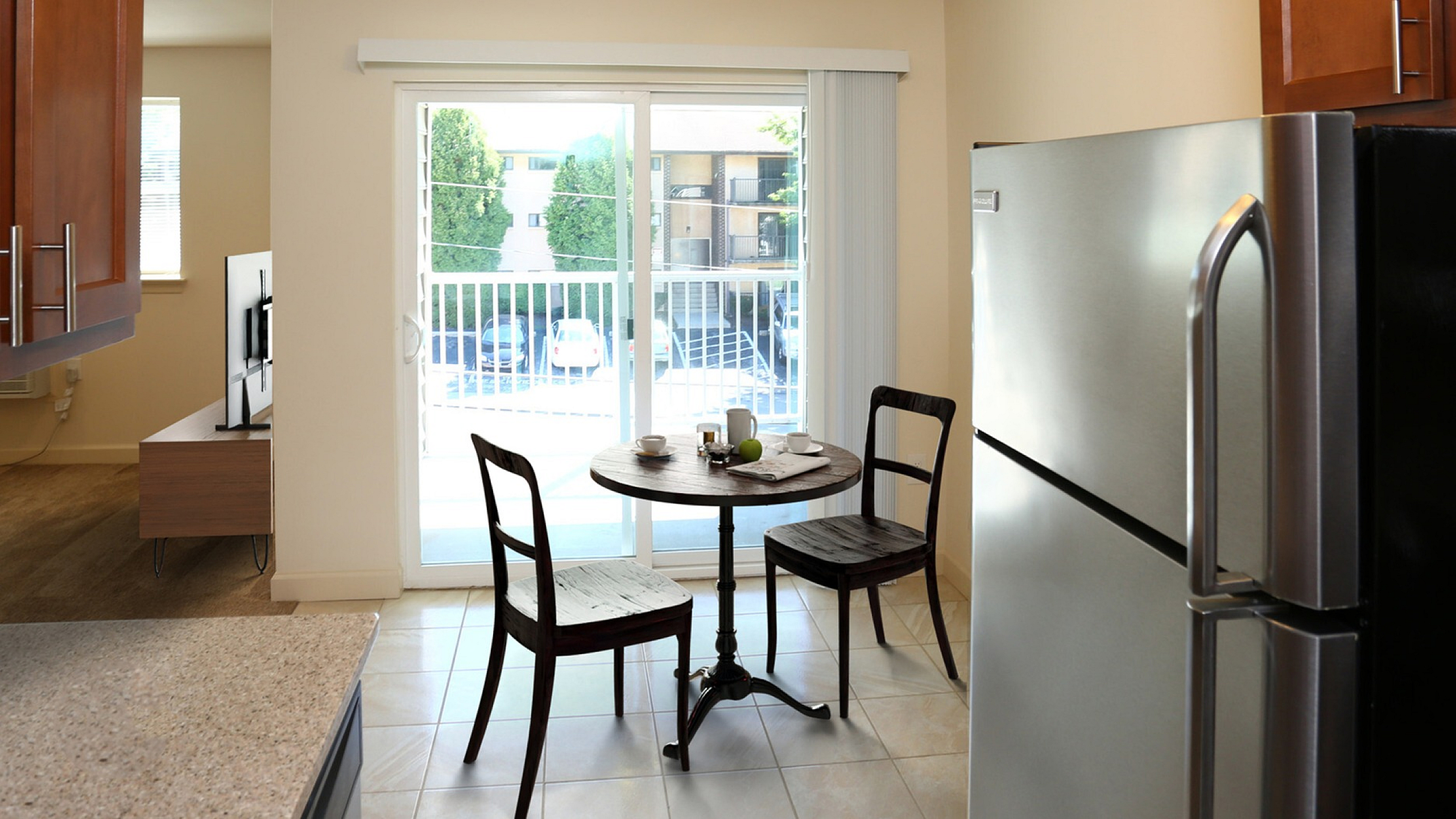 Bright and inviting kitchen area with modern refrigerator, wooden cabinets, and charming dining setup overlooking a balcony with lush greenery.