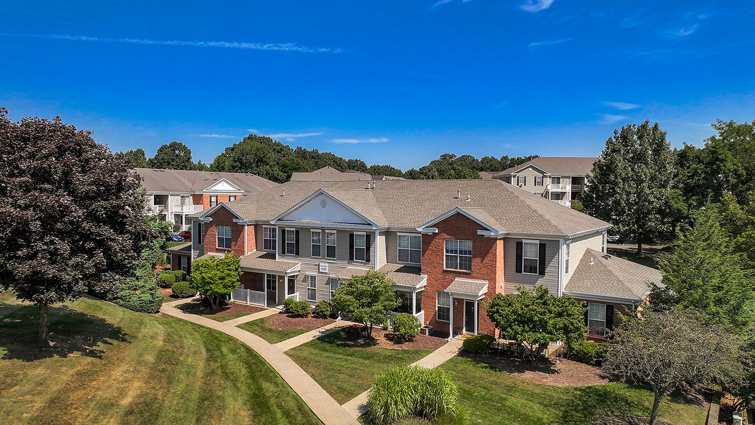 Charming brick apartment complex surrounded by lush greenery and clear blue skies, featuring well-maintained walkways and inviting entryways.