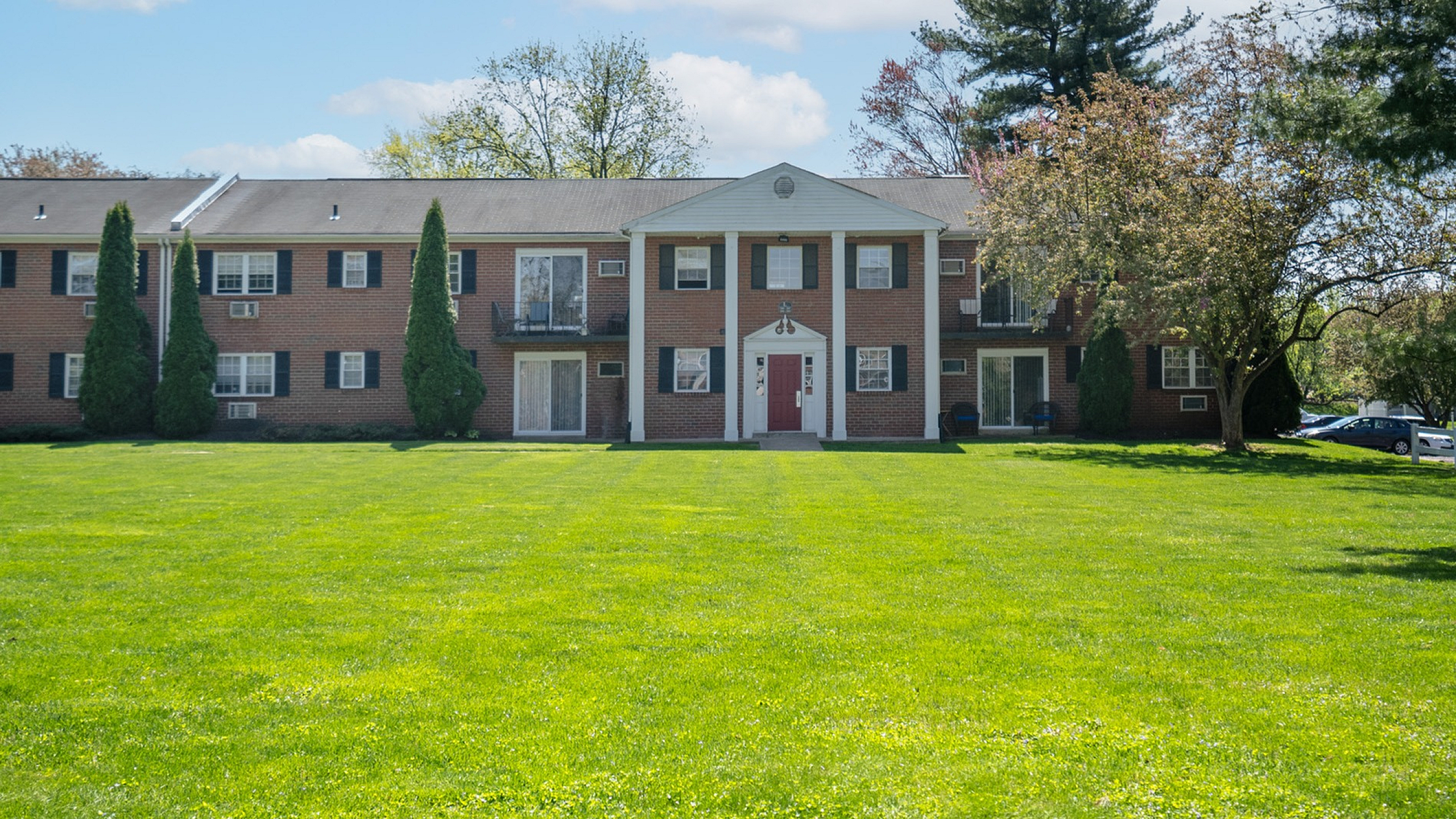 Charming two-story brick apartment building surrounded by lush green lawn and tall trees, offering a serene and inviting living environment.