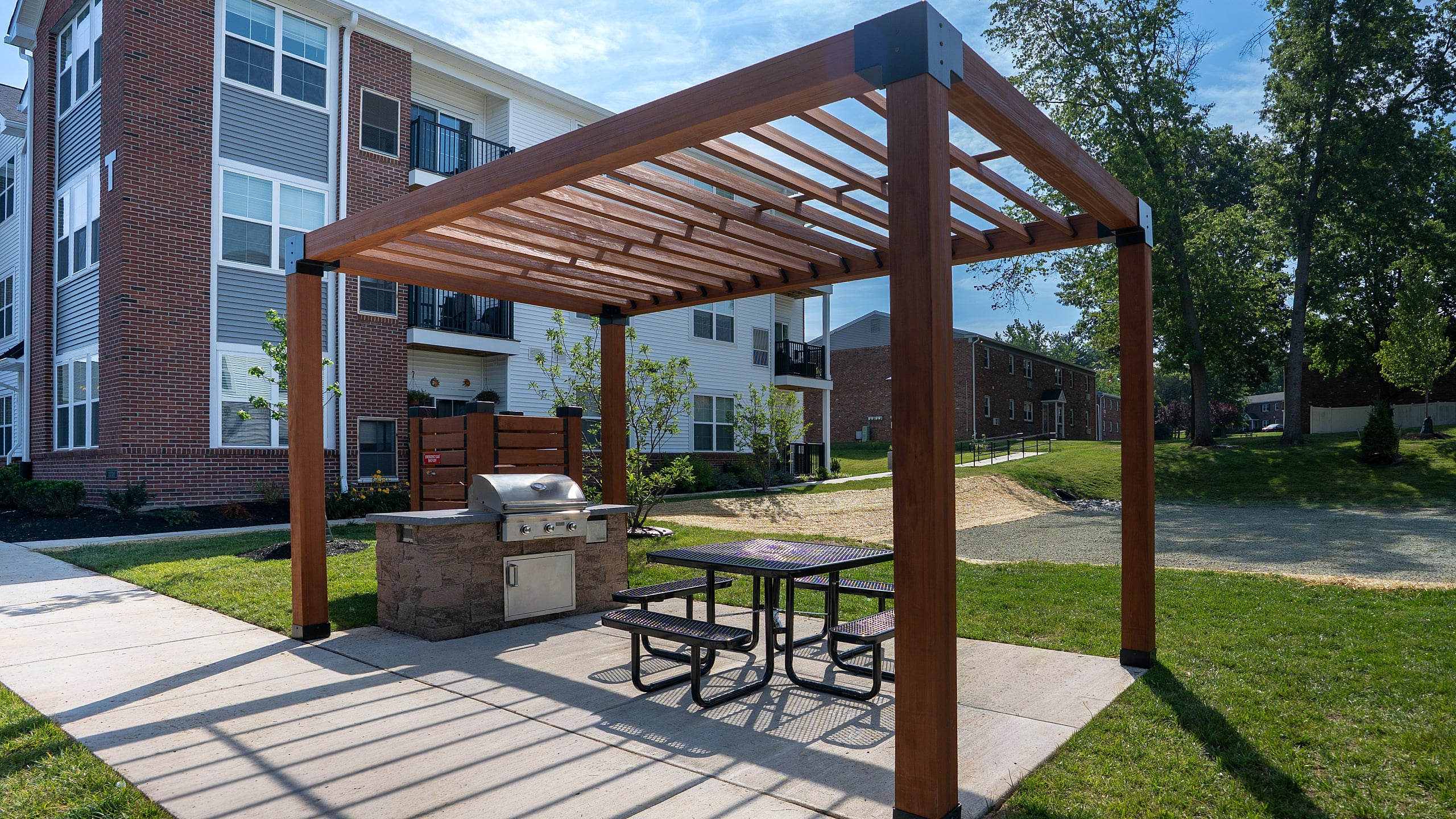 Charming apartment exterior with a stylish brick facade and a modern pergola-covered picnic area, featuring a barbecue grill for outdoor relaxation and entertainment.