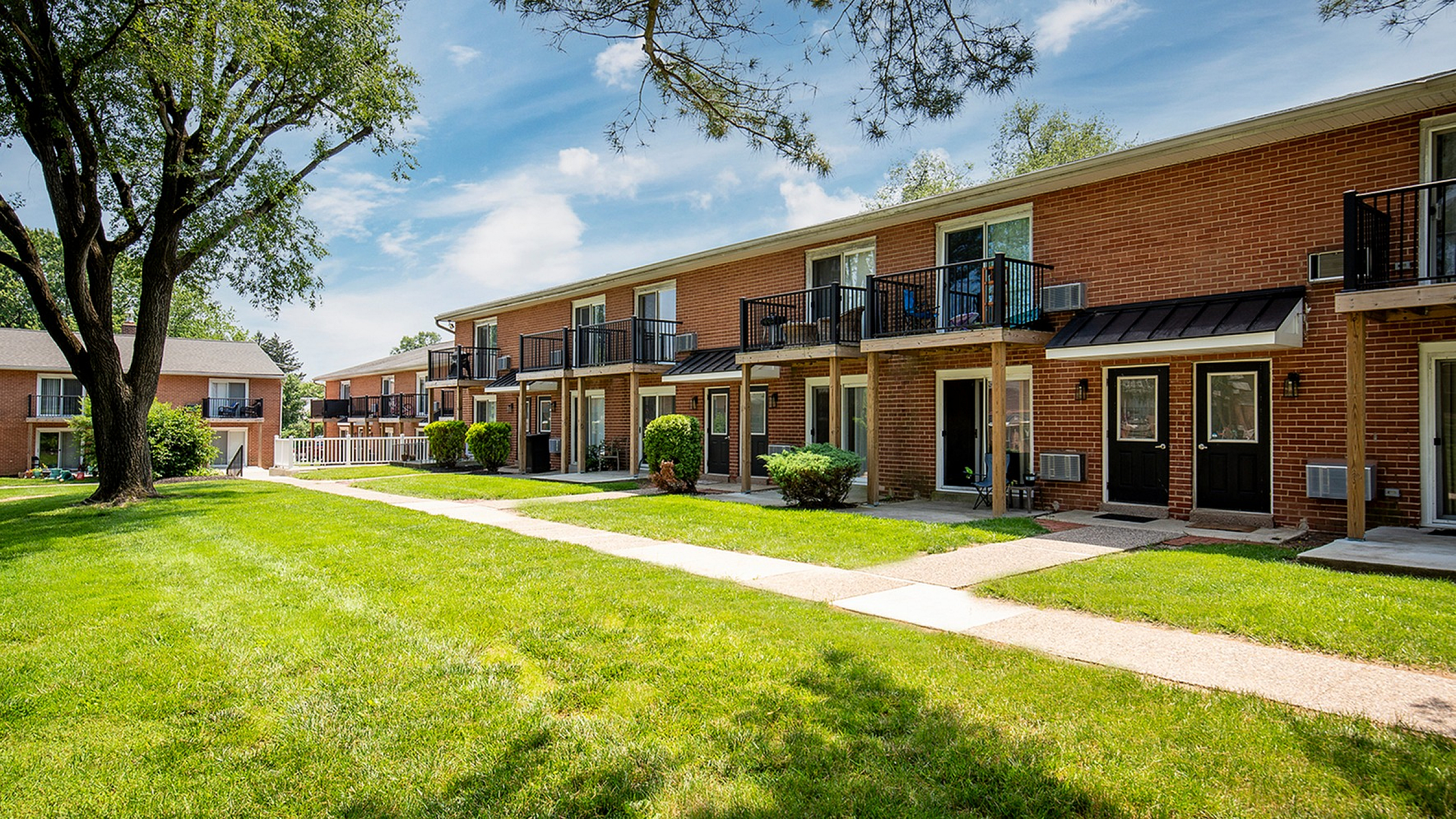 Charming brick apartment complex with private balconies overlooking lush green lawns and mature trees.