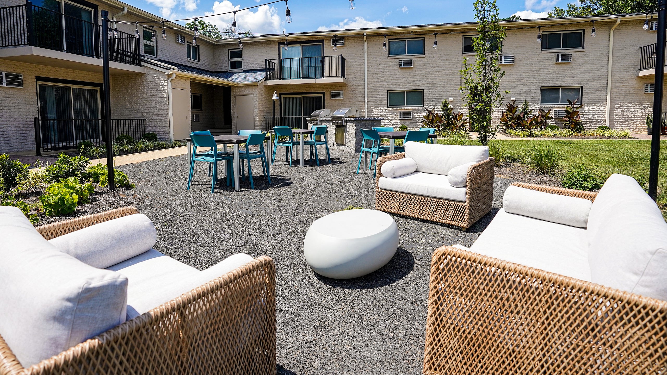 Charming courtyard view of a modern apartment complex featuring cozy outdoor seating, vibrant blue chairs, and lush landscaping under a bright blue sky.