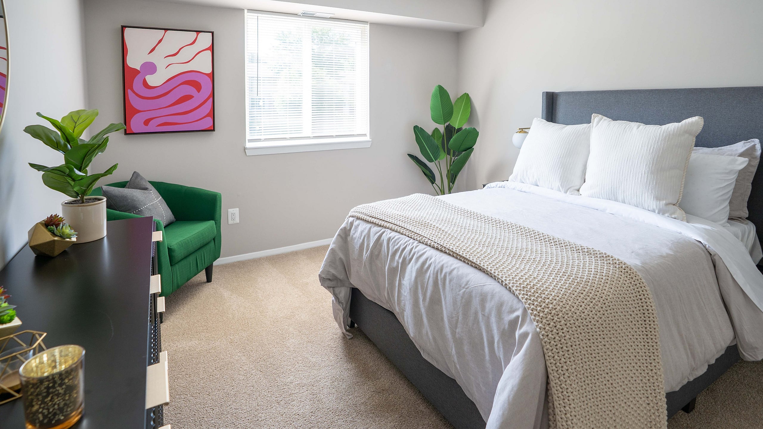 Bright and inviting bedroom with a cozy bed, elegant green armchair, and vibrant wall art, complemented by lush plants and natural light.