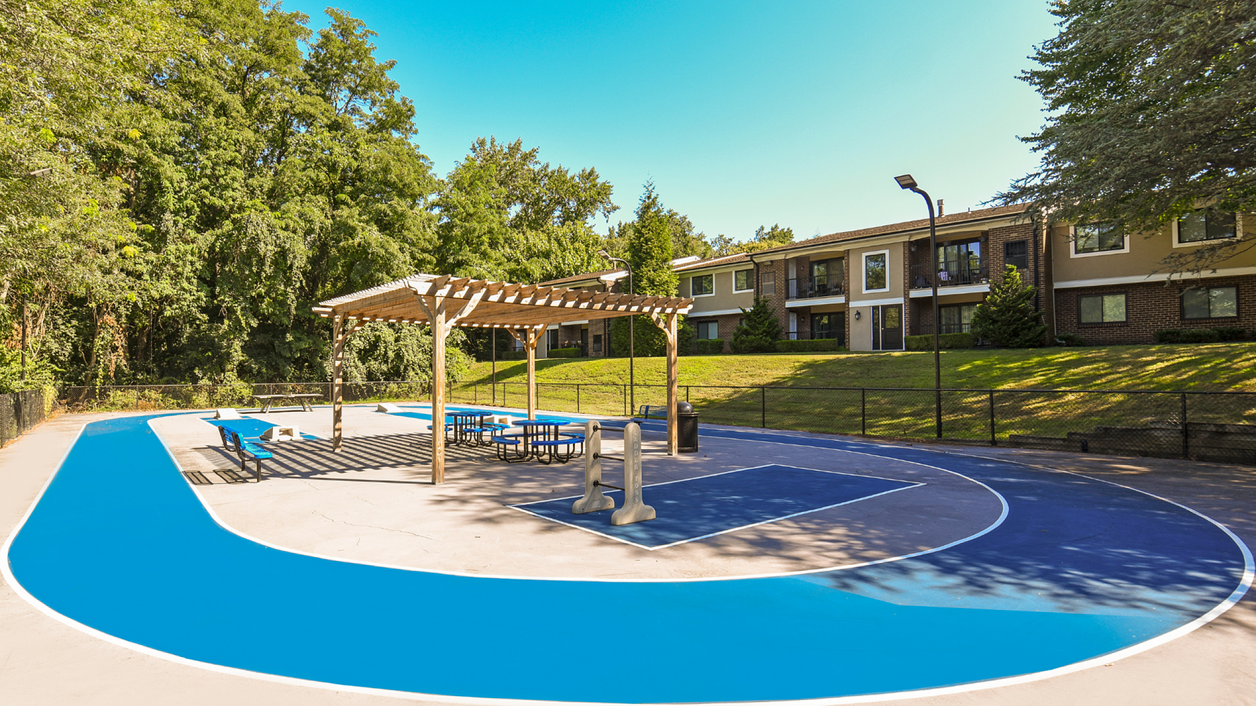 Relaxing outdoor recreational area with a vibrant blue court, pergola seating, and lush green surroundings at a modern apartment complex.