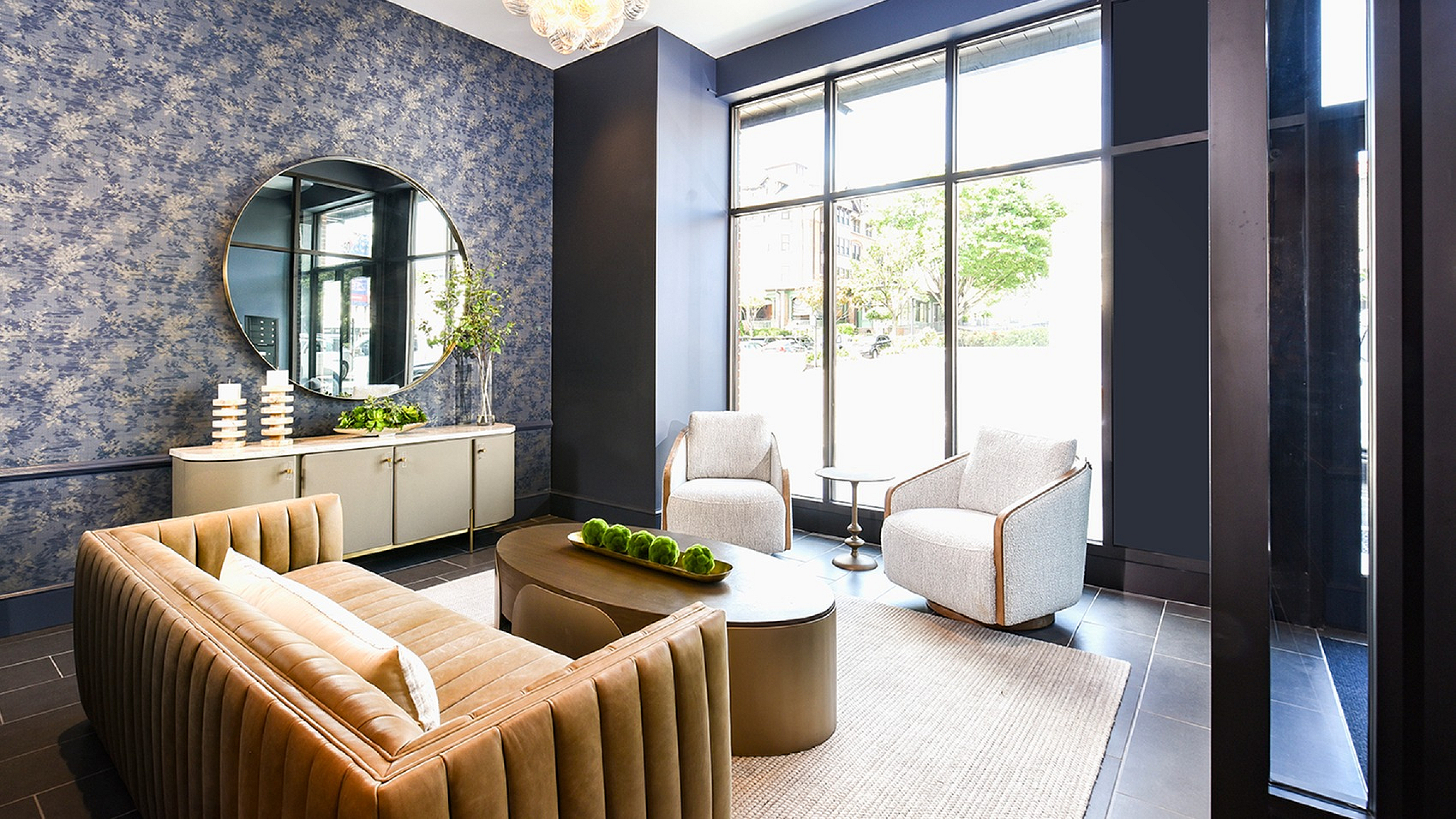 Elegant apartment lobby featuring a plush tan sofa, modern armchairs, large windows, and chic decor with accent lighting.