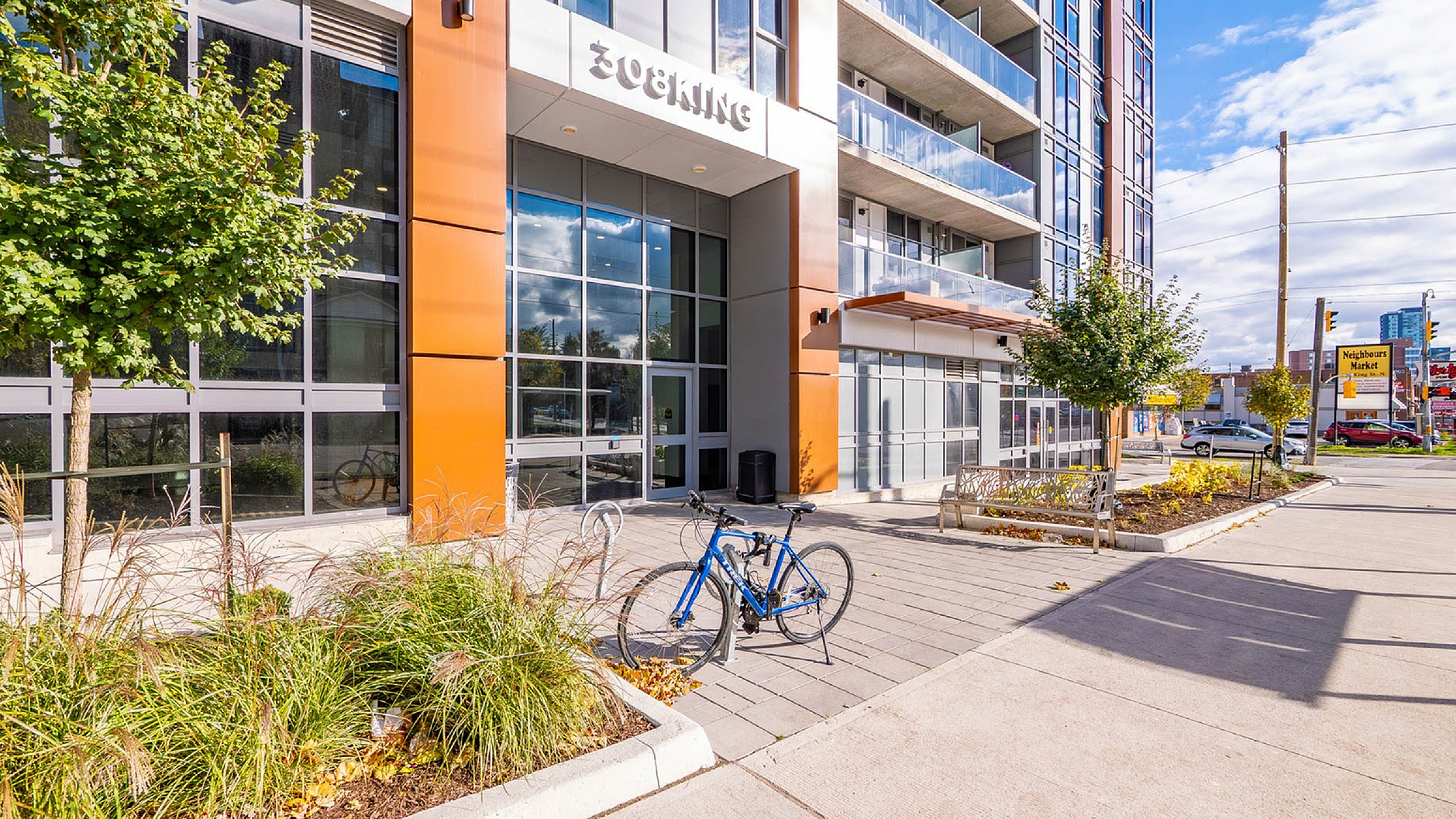 Modern apartment building entrance with sleek design, large windows, and vibrant surroundings. Features a bicycle parked on a sunlit patio, emphasizing urban lifestyle.