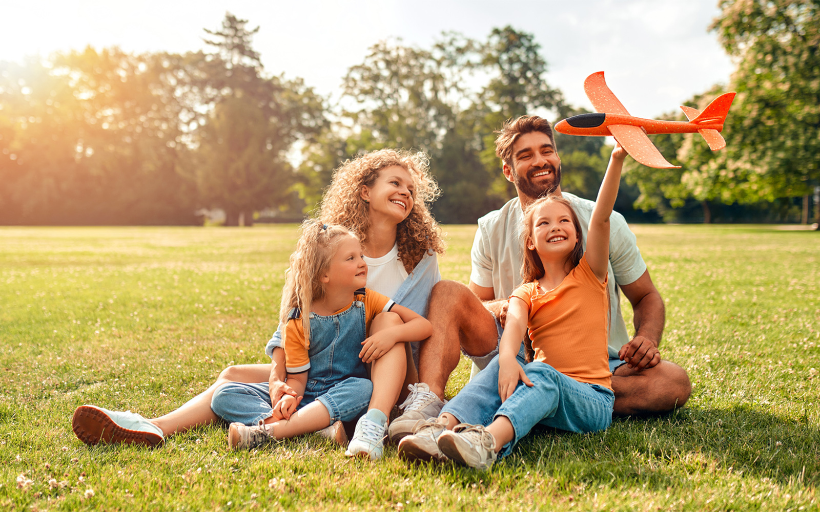 Family sitting on grass in a sunny park while a child holds up a toy airplane, with trees in the background