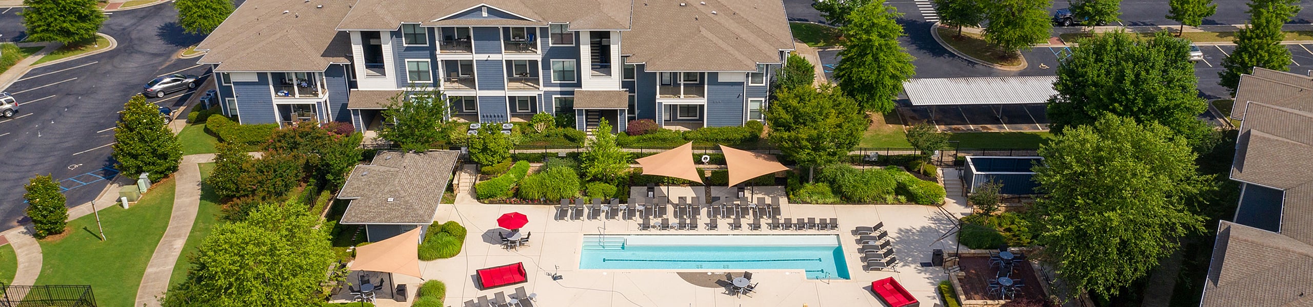 Aerial view of a modern apartment complex featuring a beautifully landscaped garden and a sparkling swimming pool with lounging chairs, shaded areas, and vibrant green surroundings.