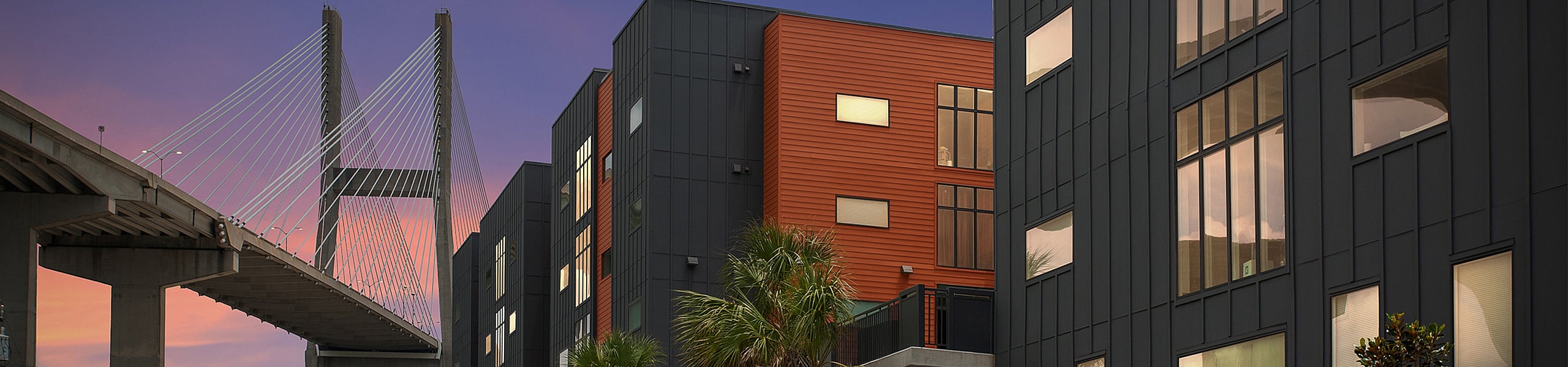 Contemporary urban apartment complex with modern architecture, adjacent to an impressive cable-stayed bridge at sunset.