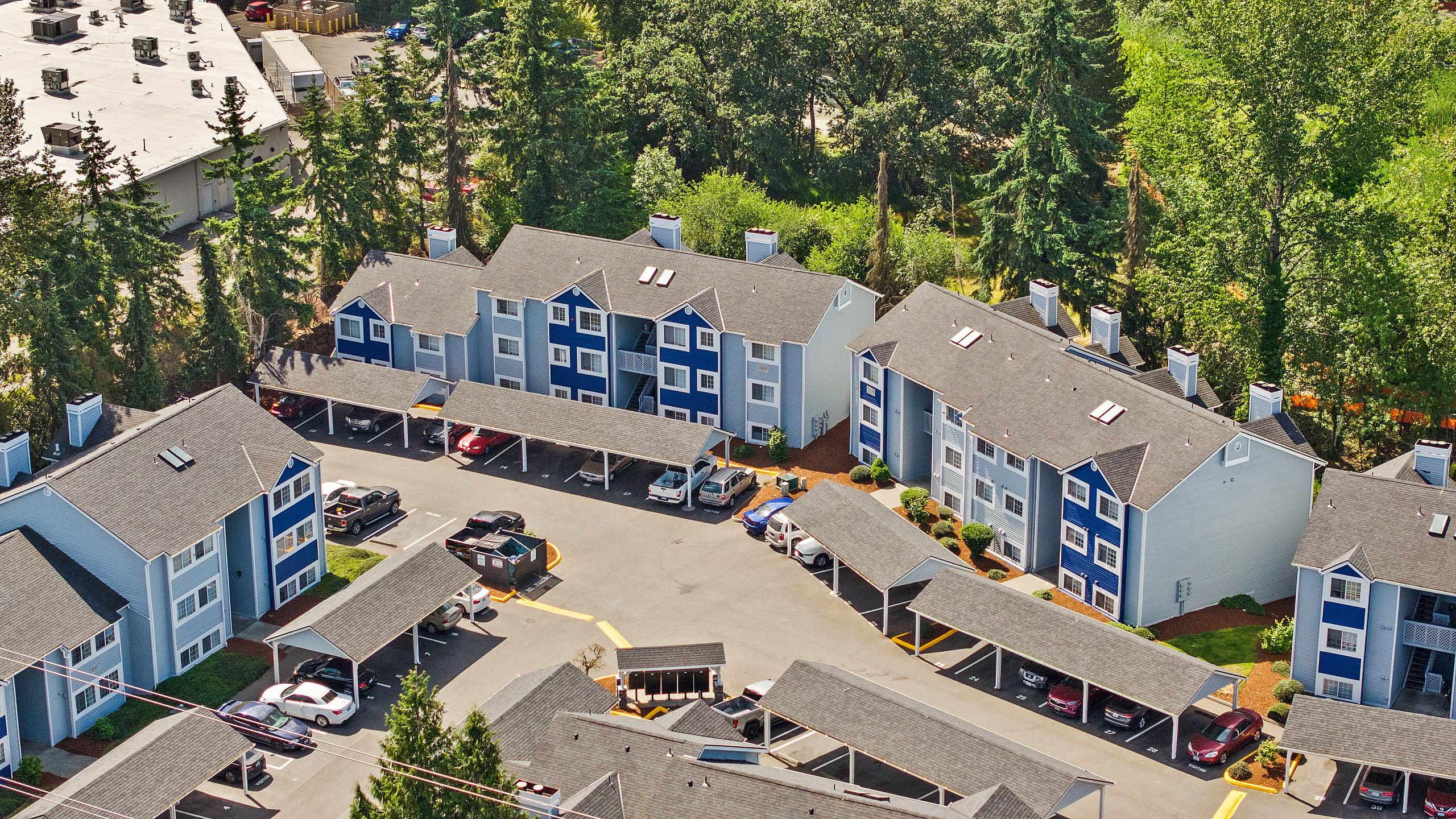 Charming aerial view of modern apartment complex surrounded by lush greenery, featuring stylish buildings with ample parking and inviting curb appeal.