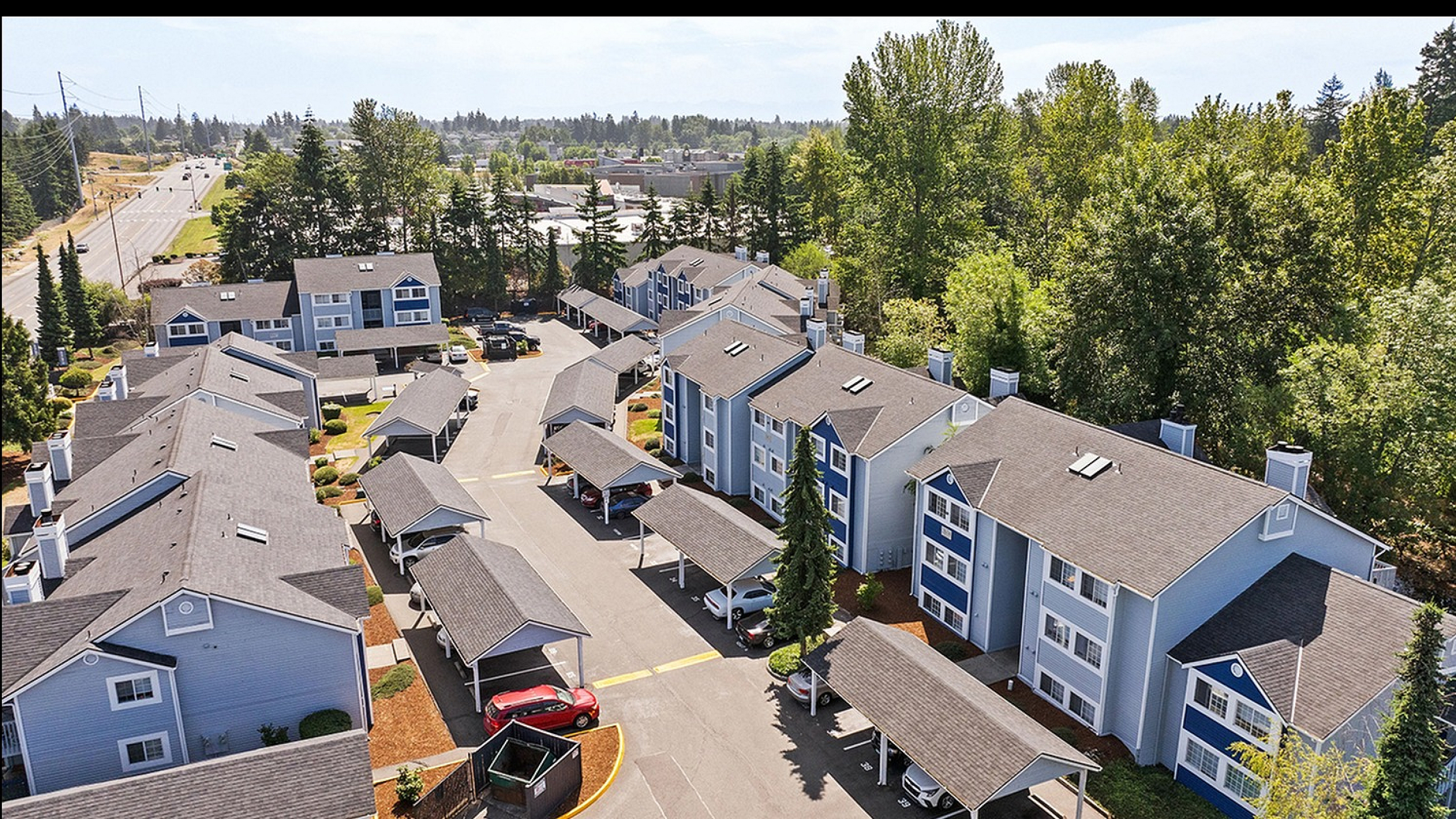 Aerial view of a charming apartment complex nestled amidst lush greenery, featuring modern blue and white buildings with covered parking, set in a tranquil suburban environment.