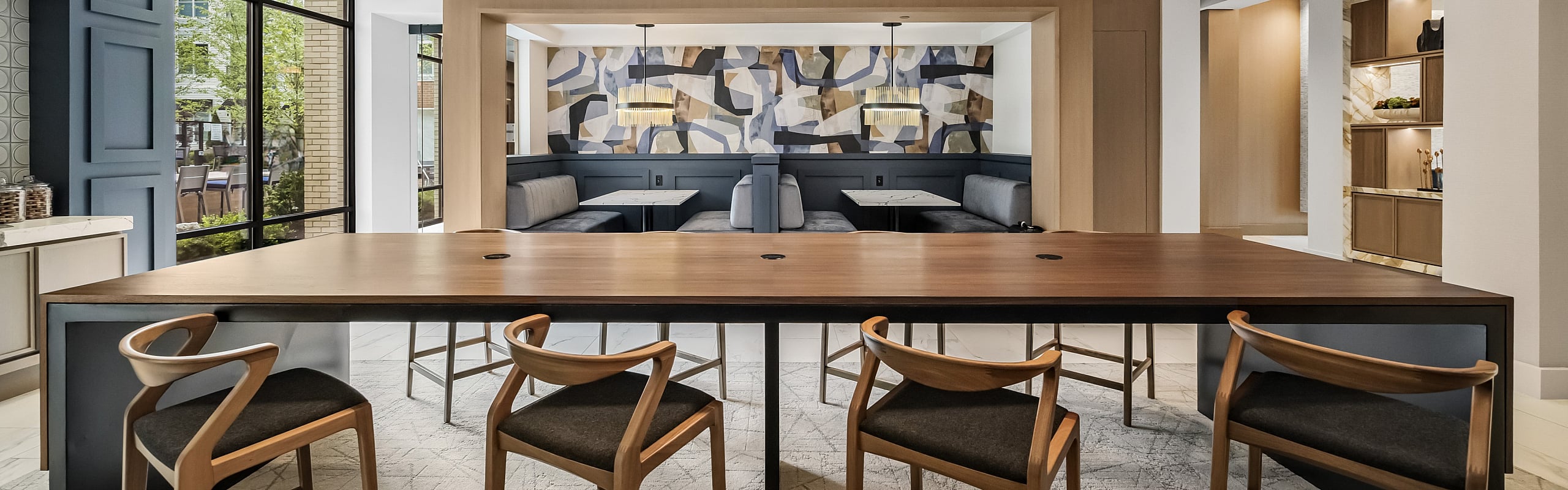 coworking lounge area with wooden 8-top communal table, pendant lighting, booth seating, abstract wall art, and large windows.