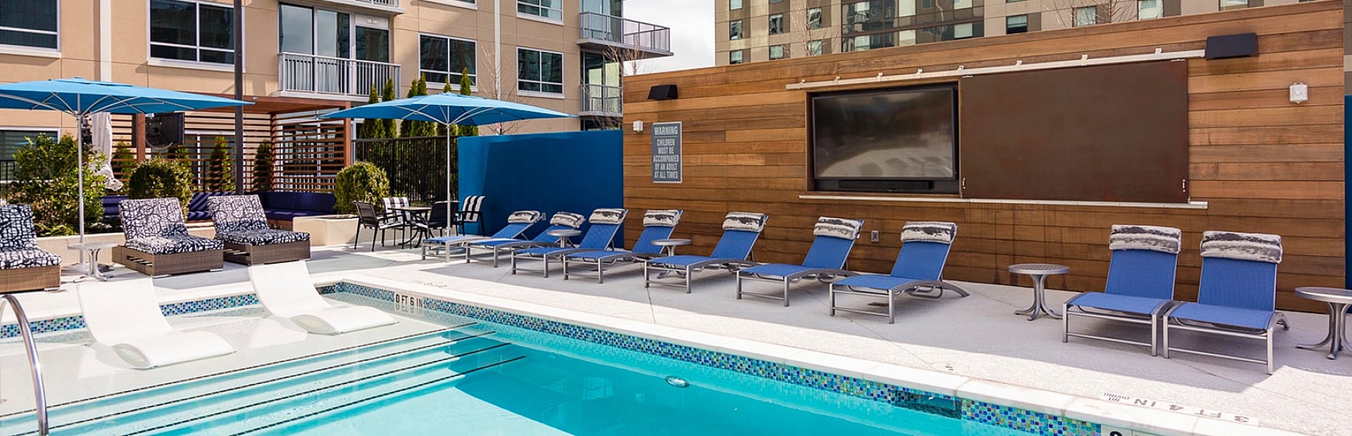 Luxurious apartment complex with a sparkling outdoor pool, surrounded by stylish lounge chairs and shaded by modern umbrellas, offering a perfect urban retreat.