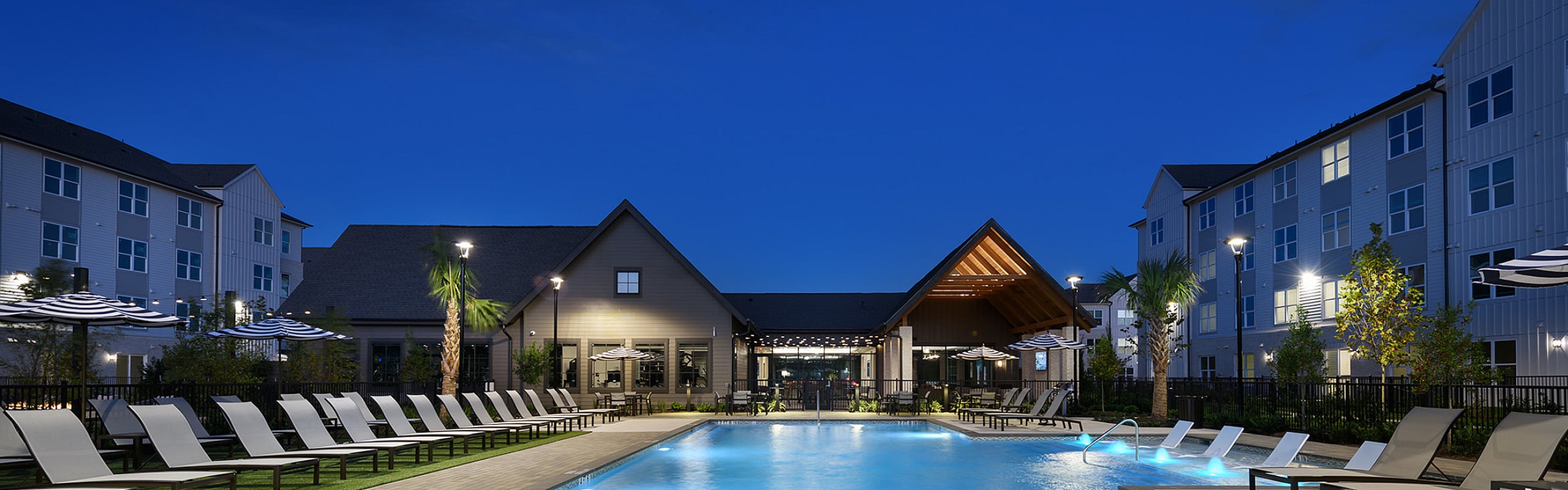 Luxurious apartment complex featuring a sparkling outdoor pool surrounded by stylish lounge chairs and modern buildings, beautifully lit against the evening sky.