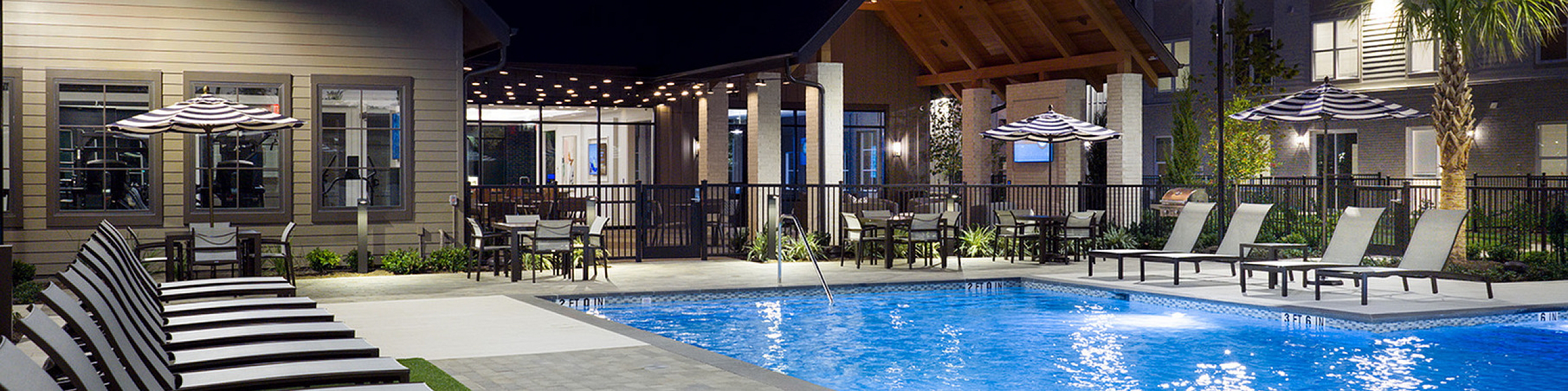 Luxurious apartment complex showcasing a beautifully illuminated pool area with sleek lounge chairs and stylish patio umbrellas, perfect for evening relaxation.