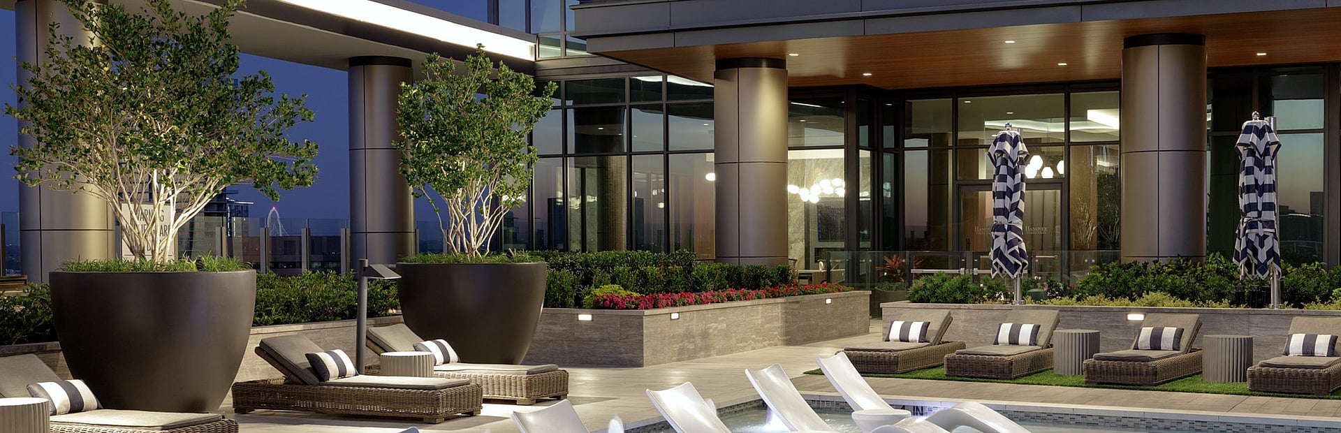 Luxurious rooftop pool area with elegant lounge chairs, surrounded by modern architecture and lush greenery, illuminated at dusk.