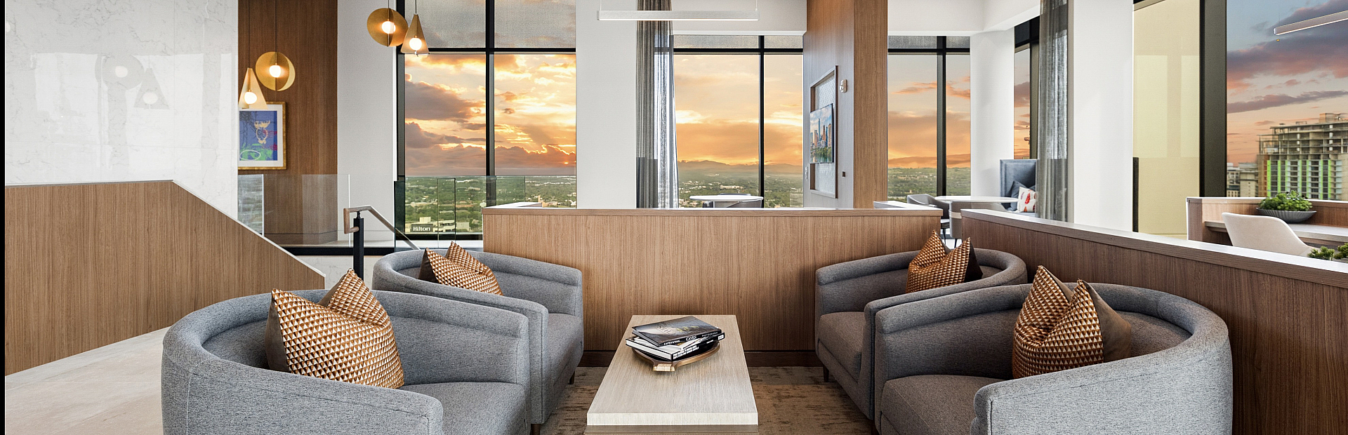 Elegant modern lounge with plush gray sofas and ambient lighting, featuring panoramic city views through expansive windows at sunset.