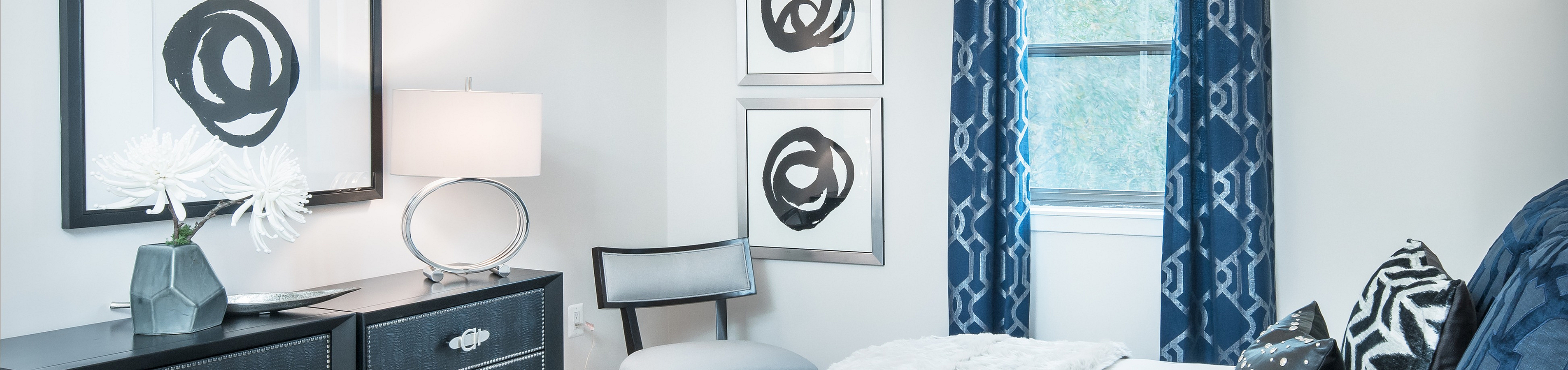 Elegant bedroom with stylish decor, featuring a plush bed with patterned pillows, chic dresser, and modern art on the walls. Natural light pours through the window, enhancing the soothing ambiance.