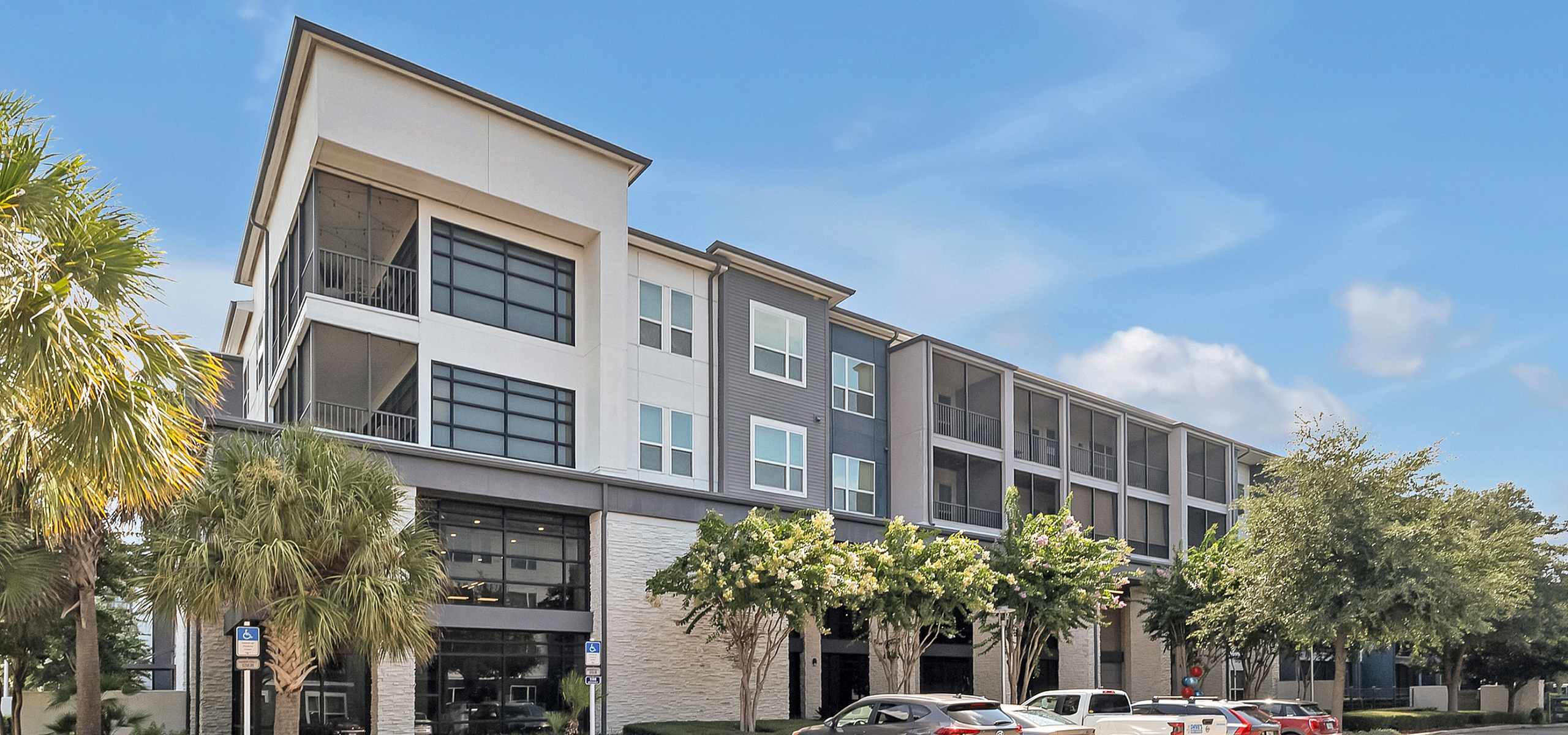 Modern apartment complex with sleek architectural design, featuring balconies and surrounded by lush greenery under a bright blue sky.